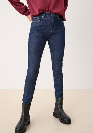 Persoon draagt donkerblauwe skinny jeans, zwarte enkellaarsjes en een bordeauxrode top met lange mouwen, staat met handen in de zij.
