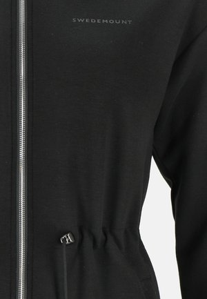 Veste zippée noire avec une texture douce, présentant une taille cintrée et des éléments en métal argenté. Logo imprimé en gris sur le côté supérieur gauche.