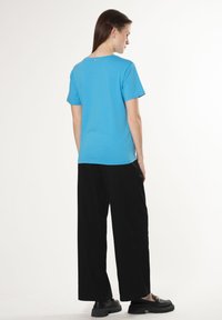 Blauwe katoenen T-shirt met korte mouwen, een ronde halslijn en een label aan de achterkant, gecombineerd met zwarte wijde broek en zwarte schoenen.