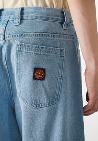 Jeans de mezclilla celeste con un parche en el bolsillo trasero que dice "Santa Cruz" en rojo y verde sobre un fondo negro.