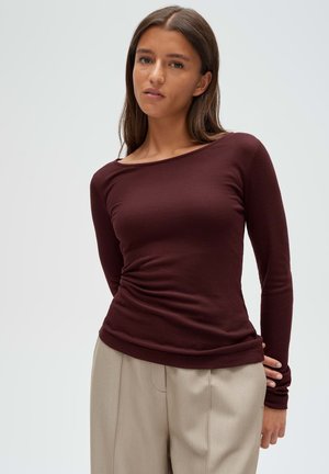 Junge Frau mit langen braunen Haaren, die ein eng anliegendes bordeauxrotes Langarmshirt und beige Hosen trägt, steht vor einem schlichten hellen Hintergrund.
