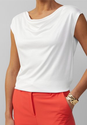 Femme portant un chemisier blanc sans manches rentré dans un pantalon rouge taille haute, avec un large bracelet doré au poignet et la main dans la poche.