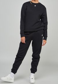 Ensemble sweat-shirt et pantalon de survêtement noir en tissu doux, avec poignets côtelés et petit logo sur la poitrine et la hanche. Baskets blanches portées.