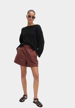 Sort oversized sweater, brune shorts med elastisk talje og sorte remsandaler. Store kvadratiske solbriller fremhæver outfittet. Enkel, afslappet stil.