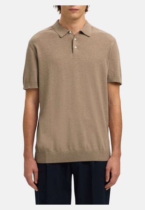 Mees, kes kannab beežikat lühikeste varrukatega kudumit polost, millel on kolm valget nööpi, koos tumedate pükstega, seisab valge tausta ees.