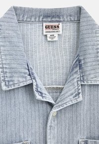 Lichtblauwe denim shirtkraag met een visgraatpatroon en zichtbaar Guess Originals merketiket en maat M/M tag.