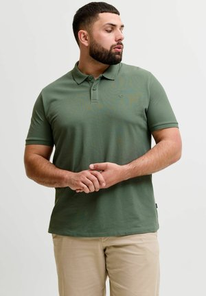 Szakállas férfi zöld galléros pólót és bézs nadrágot visel, összekulcsolt kézzel áll, oldalra néz sima háttér előtt.