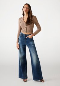 Cardigan beige a costine con scollo a V e bottoni dorati, abbinato a jeans in denim azzurro chiaro a gamba larga con dettagli strappati.