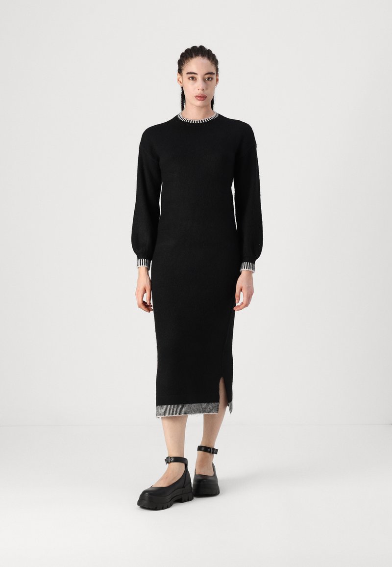 Crna, uska pletena haljina s dugim rukavima, okruglim izrezom i bočnim prorezom; ima prugasti naglasak na izrezu i rubu.