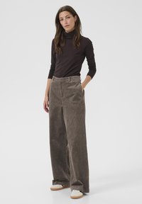 Bruine corduroy wijde broek met verticale strepen, gecombineerd met een donkere coltrui. Het model staat met één hand in de zak, draagt witte sneakers.