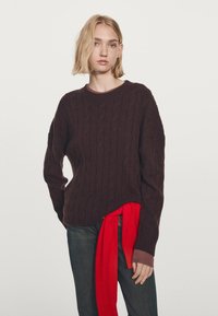 Maglione in maglia a trecce marrone con scollo rotondo, caratterizzato da un tessuto rosso a contrasto legato in vita. Indossato con jeans scuri.