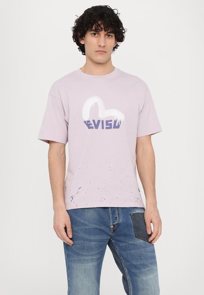 Levanduľové bavlnené tričko s bielo-tmavomodrým grafickým logom "EVISU"; má detaily striekajúcej farby na leme. Nosené s džínsovými nohavicami.