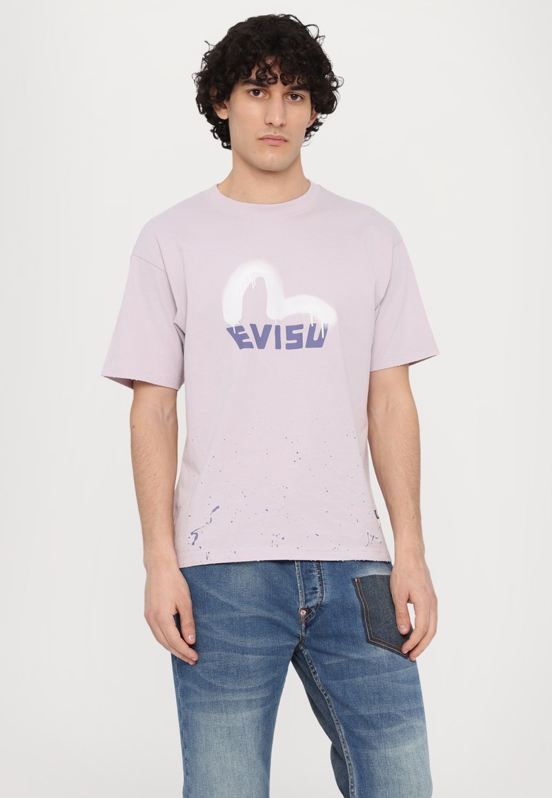 T-shirt en coton lavande avec un logo graphique blanc et bleu marine "EVISU" ; présente des détails de éclaboussures à l'ourlet. Porté avec un jean en denim.