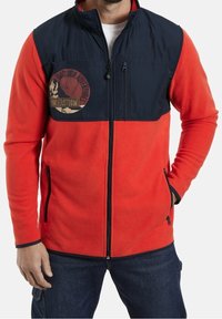 Man in een rode fleece jas met een marineblauwe schouderpaneel en ritszakken, met een logo voor buitenavonturen op de borst.