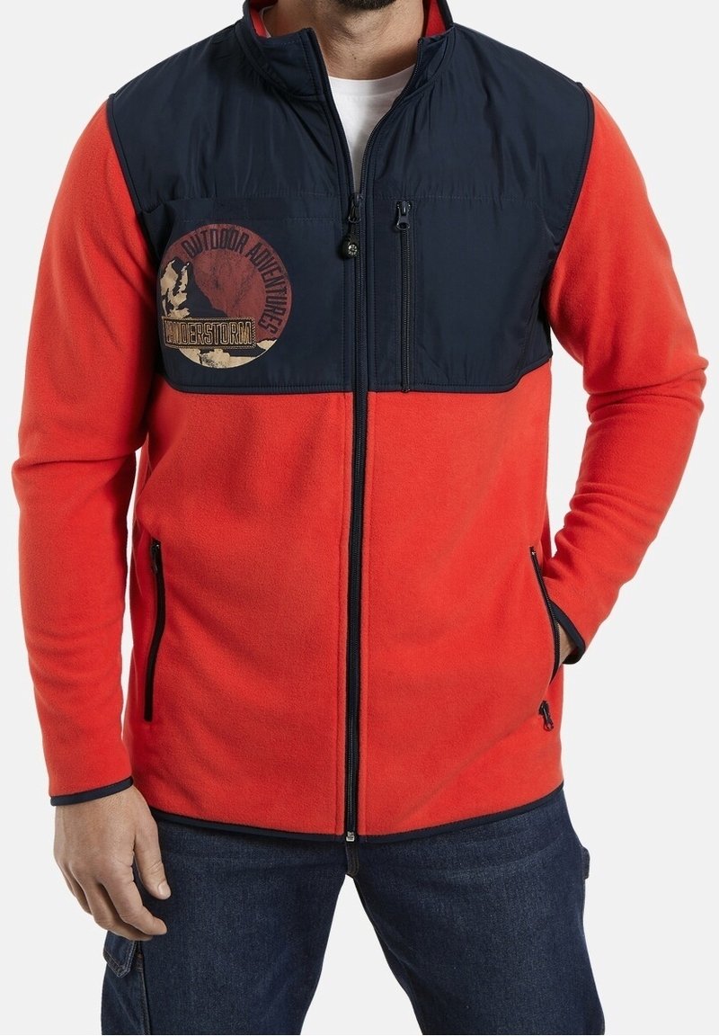 Man in een rode fleece jas met een marineblauwe schouderpaneel en ritszakken, met een logo voor buitenavonturen op de borst.