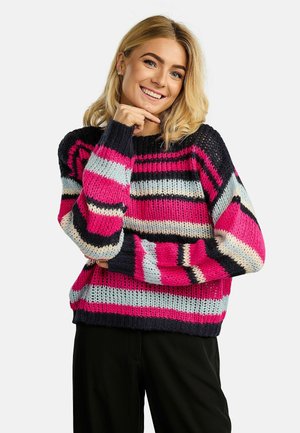Smilende kvinde med blondt hår iført en grovstrikket sweater med brede vandrette striber i pink, sort, hvid og grå, poserende med hånden nær hagen.