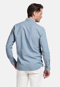 Chemise à manches longues bleu clair, avec col, en tissu texturé et avec des poignets à boutons, portée avec un pantalon crème.