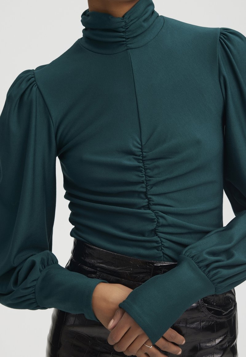 Haut col roulé turquoise froncé avec manches longues bouffantes porté avec une jupe noire texturée taille haute, mains jointes à la taille.