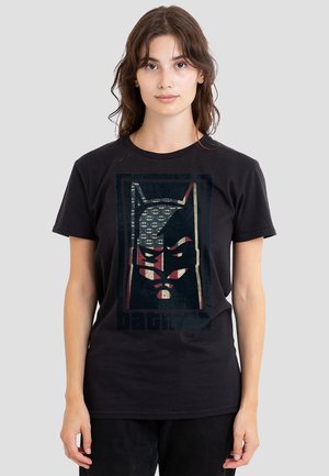 Young woman with brown hair wears black Batman graphic t-shirt featuring stylized mask and small Bat-symbol pattern on gray background.