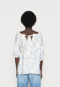 Blusa leve de creme com estampado de aquarela azul, mangas bufantes e abertura com laço nas costas. Feita de tecido suave, oferece um ajuste descontraído.