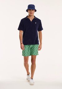 Marineblauw poloshirt, groene korte broek met palmboom patroon, witte sneakers en een blauwe bucket hat. Slim fit en casual ontwerp, gericht op zomerse kleding.