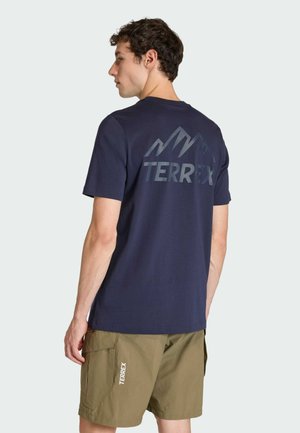 Mann trägt ein marineblaues Kurzarmhemd mit einem Berglogo und dem Schriftzug "TERREX" sowie olivgrüne Shorts mit Seitentasche und "TERREX"-Etikett.