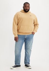 Beige hoodie with front pocket and small logo, paired with blue jeans and black canvas sneakers featuring white soles and laces.