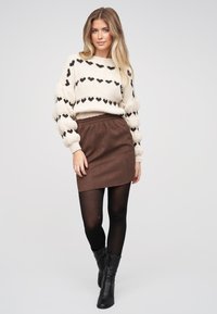 Žena v krémovém svetru s černými srdíčky, hnědé minisukni, černých punčochách a černých kotníkových botách stojící na jednobarevném pozadí.