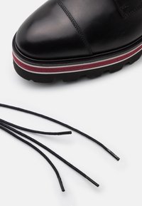 Chaussure en cuir noir à semelle épaisse avec des rayures rouges et blanches, et des lacets noirs lâches sur fond blanc.
