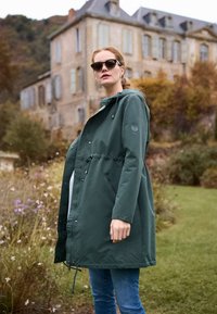 Vrouw met een donkergroene hoodie en zonnebril staat buiten bij wilde bloemen met een oud stenen gebouw en herfstbomen op de achtergrond.