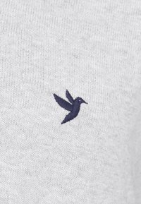 Colibri brodé bleu foncé sur un tissu tricoté gris clair, centré et montrant des coutures détaillées.