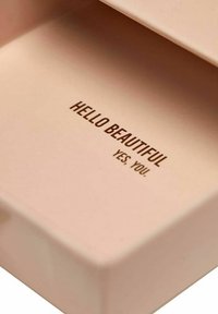 Bleknad rosa låda med tryckt text "HEJ VACKRA" och "JA. DU!" i mörkbrunt, med en slät, matt yta.