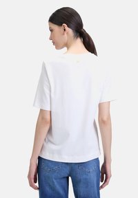 T-shirt en coton blanc avec une coupe décontractée, manches courtes, et un petit détail en forme de cœur orange près du col. Assorti à un jean en denim bleu.