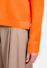 Prawa ręka spoczywa wzdłuż ciała, ubrana w pomarańczowy, puszysty sweter i beżowe plisowane spodnie na jasnym tle.