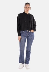 Felpa nera tagliata con cappuccio e coulisse, abbinata a jeans svasati blu e sneakers bianche. Tessuto morbido, vestibilità comoda, branding minimale.
