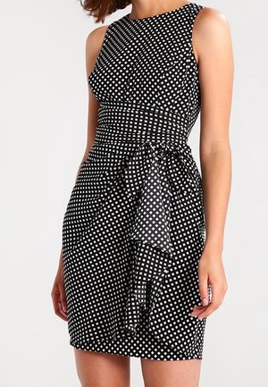 Femme portant une robe noire sans manches avec un motif carré blanc et un détail volanté asymétrique à la taille sur un fond uni.
