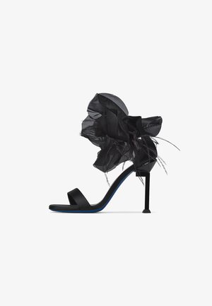 Sandalo nero con tacco alto sottile a stiletto, cinturino singolo per le dita e grande fiore decorativo in tessuto nero con piume sul cinturino alla caviglia.
