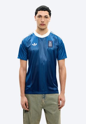 Fiatal férfi kék sportmezben, fehér gallérral és görög címerrel, világoszöld nadrágban, egyszínű háttér előtt áll.