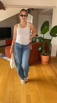 Fehér, csipkebetétes, bordázott ujjatlan top, kék, széles szárú farmernadrág, fehér, blokksarkú szandál, egy fehér kézitáska a kezében, beltéri környezet növényekkel.