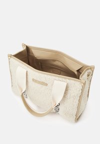 Bílá taška přes rameno z ovčí kožešiny s béžovými koženými doplňky, textilními uchy a kapsou na zip. Na přední straně je logo značky a taška má měkkou texturu.