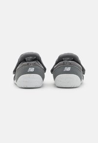 Pelēkie sporta apavi skatīti no aizmugures, ar mežģīnēm, Velcro siksnām un baltu raibumu izcilni, uz kura ir "New Balance" zīmols.