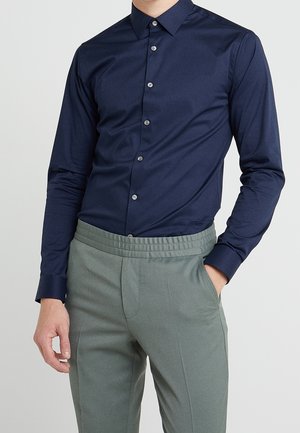 Homme portant une chemise bleu foncé ajustée boutonnée, rentrée dans un pantalon gris clair, avec une main dans la poche, devant un fond uni.