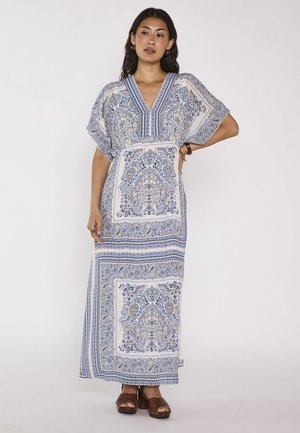 Femme debout dans une robe longue à motifs blancs et bleus, manches courtes, encolure en V et sandales marron, sur fond uni.