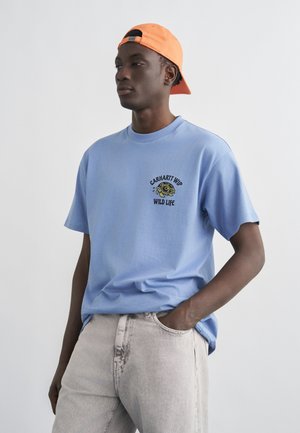 Bărbat tânăr purtând un tricou Carhartt WIP albastru deschis cu grafică cu țestoasă și o șapcă portocalie purtată invers, cu mâinile în buzunarele pantalonilor gri deschis din denim.