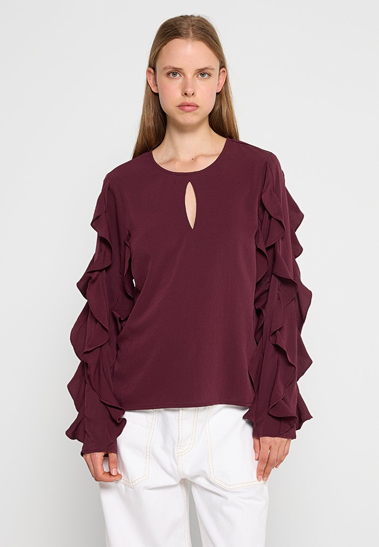 Bordeaux top met lange, gerimpelde mouwen en een sleutelgat decolleté. Gemaakt van zachte stof met een relaxte pasvorm. Gemaakt in combinatie met witte broeken.