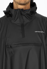 Svart vattentät pulloverjacka med hög krage, halvv zipper stängning och framficka med dragkedja. Den har en logotyp på bröstet. Slät textur.