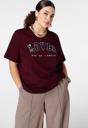 Vrouw met krullend haar in een staart draagt een bordeauxrood T-shirt met de tekst "LOVER RUE DE L'AMOUR", geruite broek, gouden ketting, creolen en armbanden.