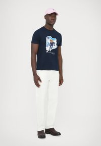 Temno modra bombažna majica s potiskom smučarja; kombinirana z belimi hlačami in črnimi čevlji; dopolnjena z roza kapo.