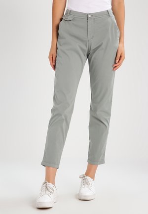 Kobieta w jasnoszarych, sięgających kostek spodniach z przodem na guziki i suwak, bocznymi kieszeniami i białych trampkach, stojąca na jednolitym tle.