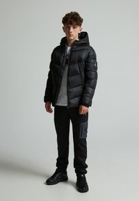 Veste noire matelassée avec capuche, présentant un design matelassé. Assortie à un pantalon noir, ajoutant une touche de poche grise. Des baskets noires complètent le look.